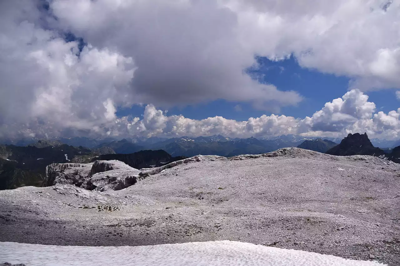 Auf dem Bild geht der Blick vom Rätschenhorn über die Gipfelhochfläche Richtung Osten. Die steinige Hochfläche nimmt einen großen Teil des Bildes ein. Im Hintergrund erkennt man rechts die dunkle Madrisa, die im Schatten liegt, und daher über den hellen Kalksteinen noch mehr auffällt. Links sieht man die Silvretta, deren Gipfel weit weg sind und zum Teil in Wolken stecken. Der blaue Himmel schaut nur an wenigen Stellen heraus, die Quellwolken sind zum Teil sehr dicht und werfen viele Schatten.