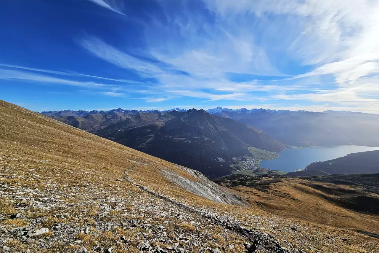 Auf dem Bild sieht man im Vordergrund den Weg im Schlussabschnitt des Anstiegs zum Piz Lad. In diesem Bereich beschreibt der Weg eine weit ausholende Kehre. Im Hintergrund erkennt man die Gipfel der Ötztaler Alpen, aus denen Glockturm, Weißseespitze, Plamorder Spitze und Weißkugel herausragen. Rechts im Bild sieht man den Reschensee mit der gleichnamigen Ortschaft. Der Himmel ist blau, es hat viele Schleierwolken, die den Himmel verzieren.