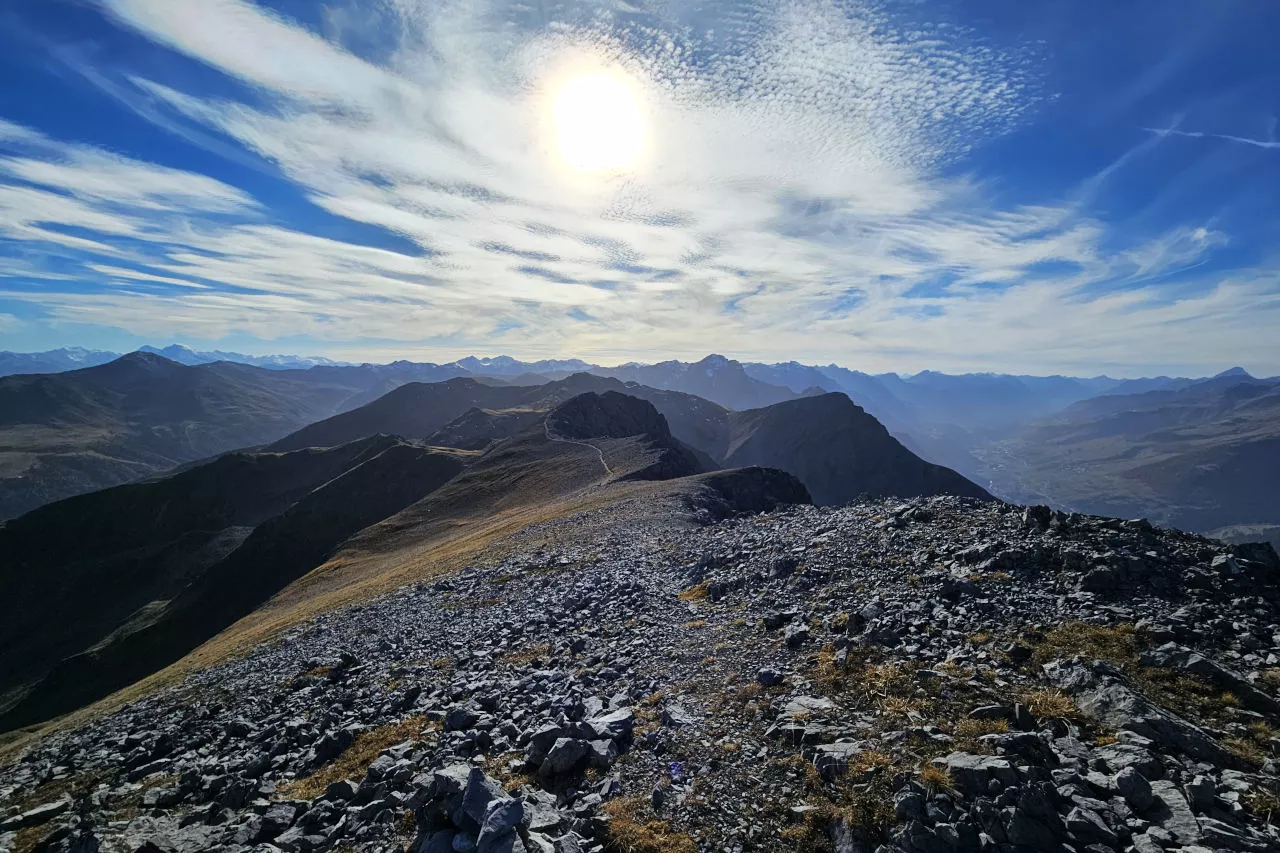 Auf dem Bild geht der Blick vom höchsten Punkt des Piz Lad Richtung Südwesten zu den Gipfeln der Unterengadiner Dolomiten. Links sieht man im Hintergrund noch die Ortlergruppe, rechts am Bildrand den Piz Linard in der Silvretta und das Unterengadin mit seinen Dörfern. Im Mittelgrund erkennt man den dritthöchsten Punkt des Piz Lad und den Weg, der auf ihn führt. Im Vordergrund erkennt man Kalkgeröll auf dem flachen Gipfel. Der Himmel ist blau, es hat viele Schleierwolken, die den Himmel verzieren und diese Gegenlichtaufnahme möglich machen - sie decken die Sonne zum Teil ab.