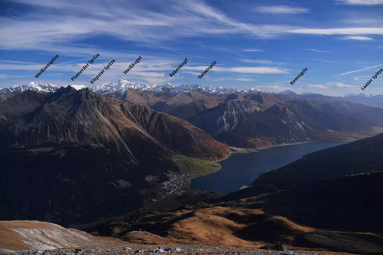 Auf dem Bild geht der Blick vom Piz Lad zu den Gipfeln der Ötztaler Alpen zwischen Weißseespitze und Remsspitz. Die bedeutendsten Gipfel sind auf dem Bild beschriftet. Die Gipfel über 3000 m sind bereits verschneit. Darunter liegt nur in den schattigen Nordflanken etwas Schnee. Die Landschaft wirkt sehr herb. Daran ändert auch der blaue Reschensee nichts. Er ist in der rechten Bildhälfte gemeinsam mit der gleichnamigen Ortschaft zu sehen. Rechts am Bildrand erkennt man noch die Laaser Spitze in der Ortlergruppe. Im Vordergrund sieht man Teile der Südflanke des Piz Lad, durch die der Anstieg verläuft. Der Himmel ist blau mit Schleierwolken.