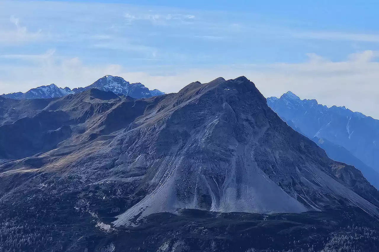 Auf dem Bild geht der Blick von Ebene oberhalb Nauders zum Piz Lad, der sich knapp rechts der Bildmitte erhebt. Nach dieser Seite zeigt er eine steile Felswand aus Kalk und auch nach rechts (Norden) fällt der Gipfel fast senkrecht ab. Nach links (Süden) erkennt man die weniger steile Seite, über die der Anstieg verläuft. Man sieht auch sehr schön die drei Gipfelpunkte. Im Hintergrund zeigen sich links Piz S-chalambert Dadaint und Piz Pisoc rechts, zwei 3000er in den Unterengadiner Dolomiten. Der Himmel ist blau mit vielen Schleierwolken.