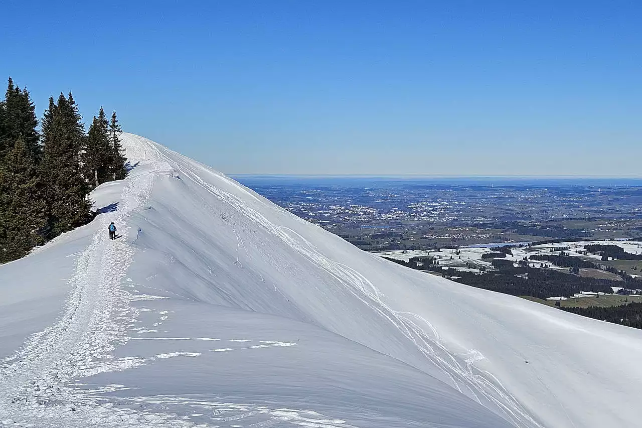 Auf dem Bild sieht man zwei Winterwanderer auf dem einfachen und breiten Grat zum Wertacher Hörnle Hauptgipfel. Sie befinden sich gerade in der Mitte zwischen dem Vorgipfel und dem Zwischengipfel, den man links oben sieht. Während das Gelände am Wertacher Hörnle noch tief verschneit ist, liegt im Alpenvorland rechts im Hintergrund kaum Schnee. Der Himmel ist blau und wolkenlos. Es ist ein außergewöhnlich klarer Tag.