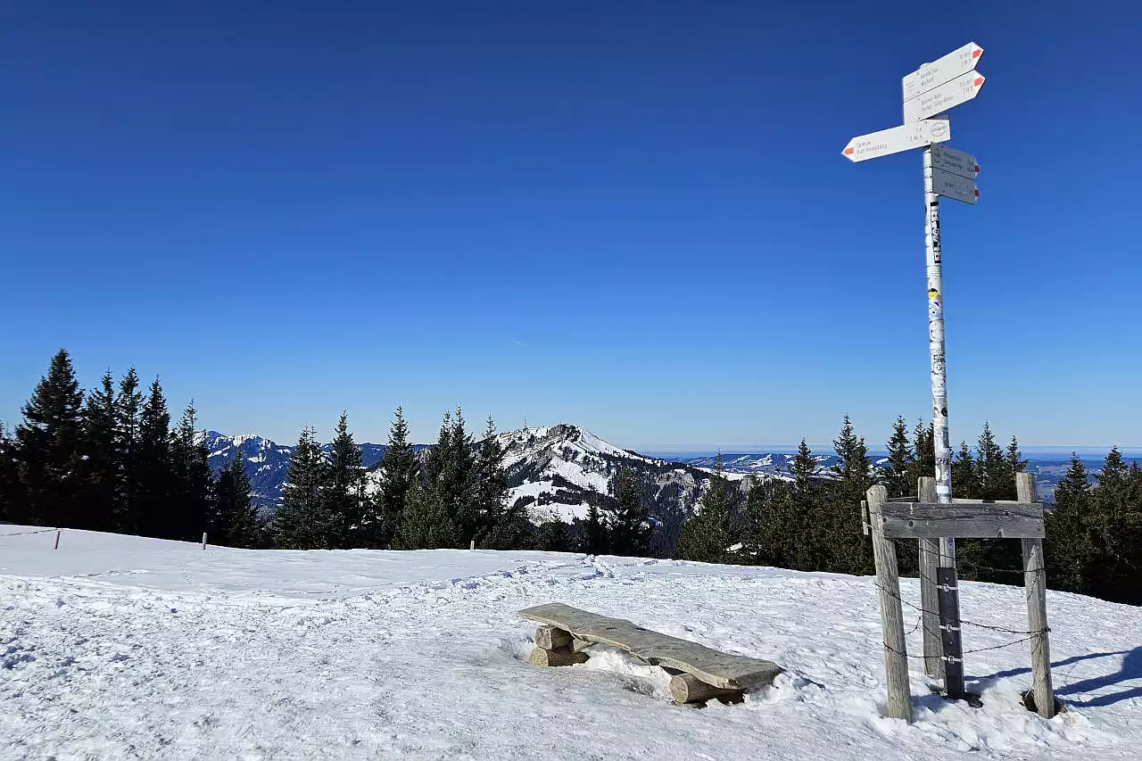 Auf dem Bild sieht man den Wegweiser und die Ruhebank am Wertacher Hörnle Vorgipfel. Hier liegt noch viel Schnee. Im Hintergrund sieht man den Grünten und rechts davon den flachen Hauchenberg. Links vom Grünten sieht man noch die ersten Gipfel der Nagelfluhkette, die anderen Gipfel sind durch Bäume verdeckt. Der Himmel ist blau und wolkenlos. Ein wunderbarer Wintertag.