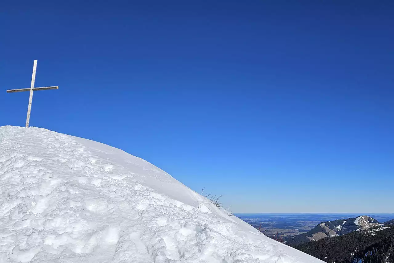 Auf dem Bild sieht man links oben das schlichte Gipfelkreuz am Wertacher Hörnle. Es scheint aus einem riesigen Schneehaufen herauszuragen. Im Schnee erkennt man auch die Spuren der Winterwanderer und Schneeschuhwanderer. Rechts geht der Blick über den Gipfel der Reuterwanne hinaus ins Alpenvorland. Dort liegt kaum Schnee. Der Himmel ist blau und wolkenlos. Es ist ein außergewöhnlich klarer Tag.