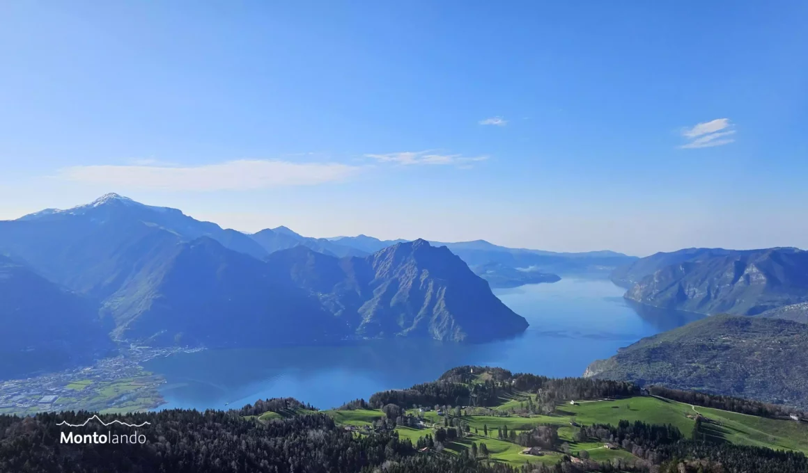 Auf dem Bild geht der Blick vom Monte Colombina Richtung Süden zum Iseosee, den man von hier fast ganz überblicken kann. Links vom See sieht man die markanten Gipfel des Monte Guglielmo links und der Corna Trentapassi in der Bildmitte. Im Vordergrund sieht man in der Tiefe die Weiden und Wälder auf der Hochfläche der Ortschaft Bossico. Der Himmel ist blau, es hat ganz wenige Schleierwolken.