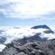 Auf dem Bild geht der Blick vom Signal du Petit Mont Cenis Richtung Süden über die Gegend um den Col du Mont Cenis hinweg zu den Gipfeln des Rocciamelone und des Mont Giusalet. Über der Piemont-Ebene liegt ein dichtes Wolkenmeer, die Wolken ziehen auch um die Gipfel. Der Himmel ist blau mit zum Teil dichten Schleierwolken.